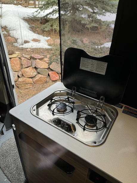 RV rental: Open two-burner gas stovetop in a camper kitchen, offering a wintery view of snow-covered rocks & trees.