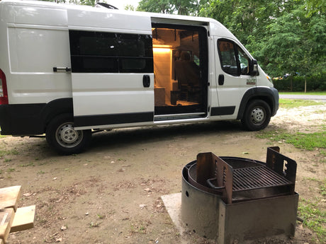 White RV rental camper van with open door and lighted interior at a dirt campsite, with a metal fire pit in the foreground.