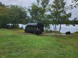 Black RV rental van parked lakeside on a grassy campsite with trees, picnic table, & fire pit under an overcast sky.