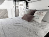 Inside an RV rental: a made bed with an "Adventure Awaits" pillow, white paneled walls, and open back doors.
