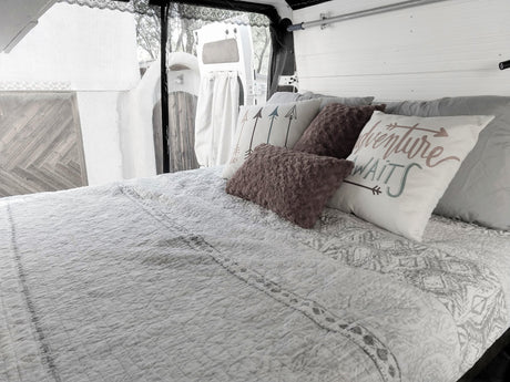 Inside an RV rental: a made bed with an "Adventure Awaits" pillow, white paneled walls, and open back doors.