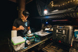 Nighttime RV rental setup: Man cooking outdoors with portable kitchen, fridge, and rooftop tent.