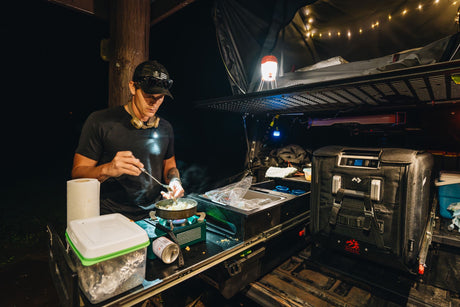 Nighttime RV rental setup: Man cooking outdoors with portable kitchen, fridge, and rooftop tent.