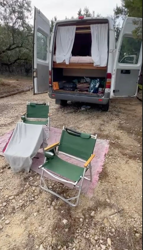 Cozy rv rental setup. Open camper van doors show a bed, storage. Two chairs on a rug invite outdoor relaxation.