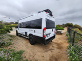 This white Tellaro RV rental is parked on a dirt lot by a picnic table and a fence amidst green plants under a cloudy sky.