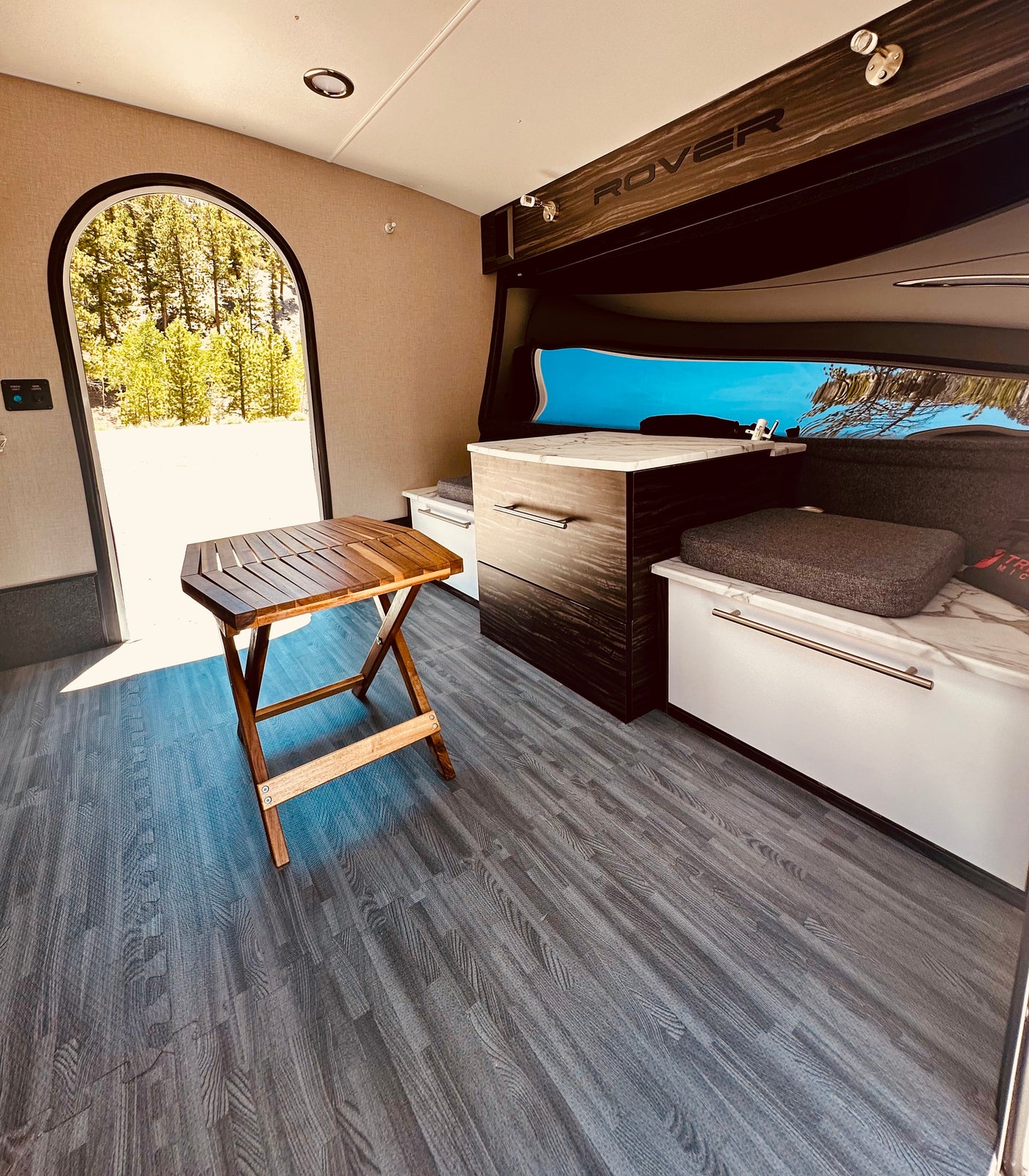 Inside a cozy RV rental: features a wooden table, modular floor, and archway to nature. Perfect for glamping.