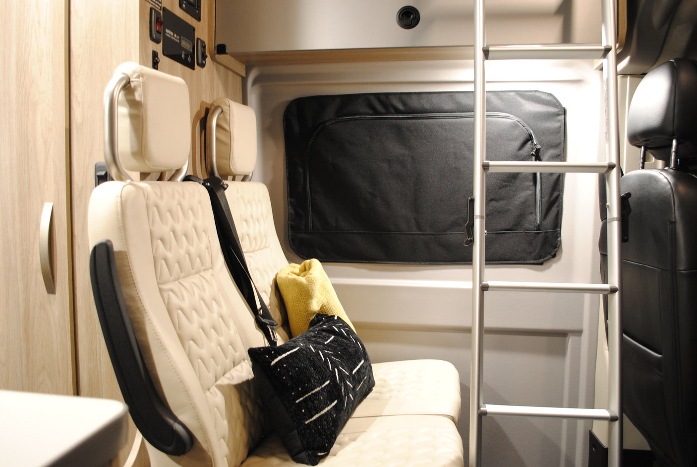 RV rental interior featuring cream passenger seats, control panel, black window covers, and a ladder to an upper bunk.