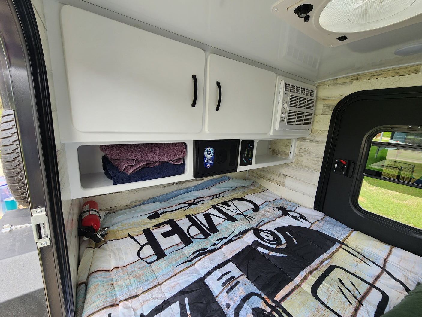 RV rental bedroom view: bed with 'CAMPER' comforter, wood walls, overhead cabinets, AC, and a security box.