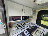 RV rental bedroom view: bed with 'CAMPER' comforter, wood walls, overhead cabinets, AC, and a security box.