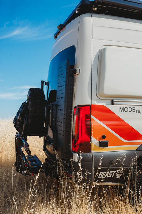 Experience overlanding with this 4x4 rv rental van, featuring custom graphics, rear gear panel, & spare tire in a field.