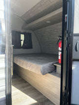 Cozy RV rental interior: bed, fire extinguisher, and wood-paneled walls inside a compact travel trailer.