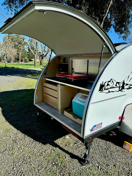 View of a teardrop RV rental's open rear kitchen, showing a red stove, cooler, and wooden drawers for compact camping.