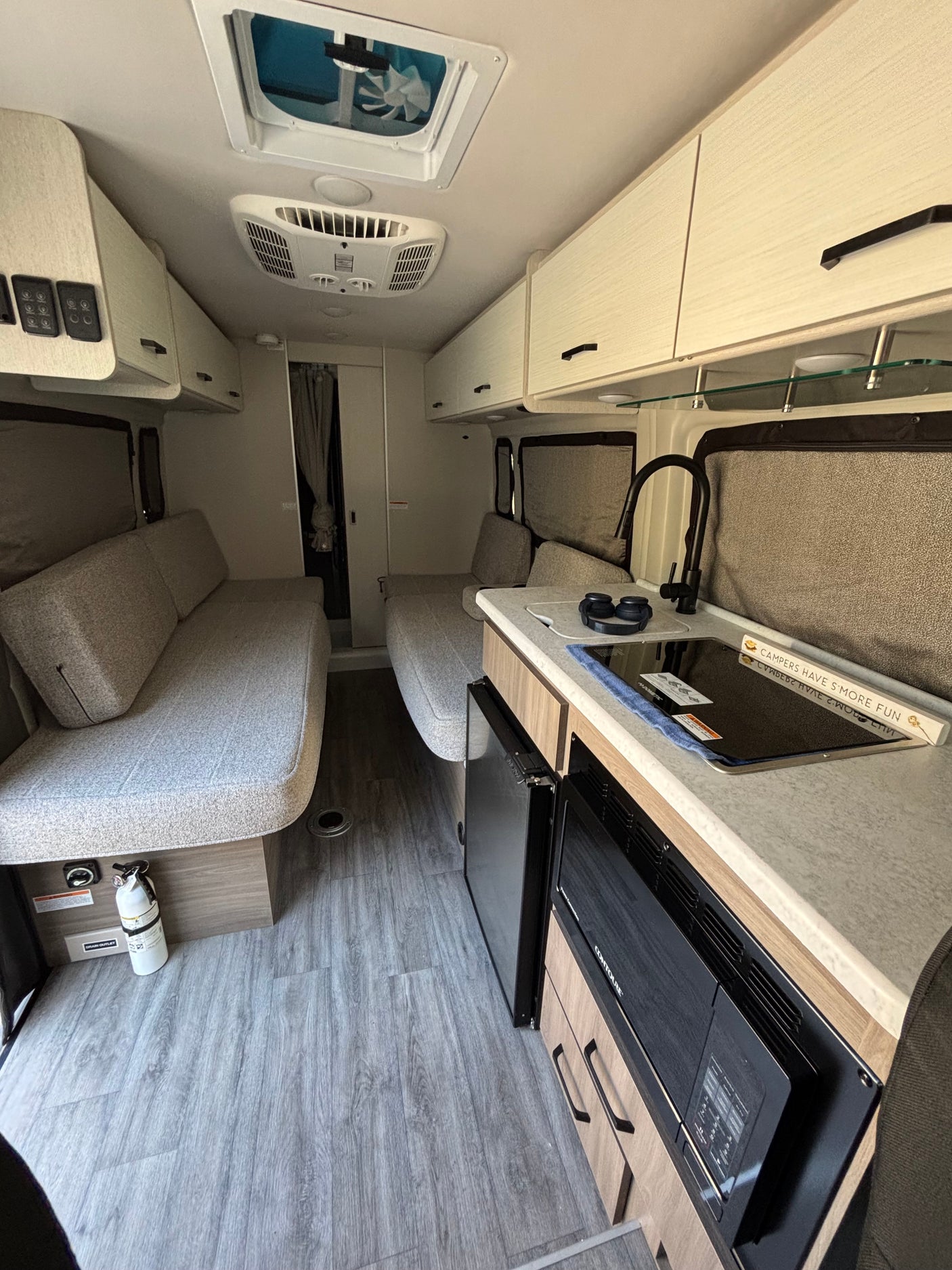 Modern rv rental interior: kitchenette with sink, stovetop, microwave & fridge, plus comfortable seating, overhead storage, and AC.