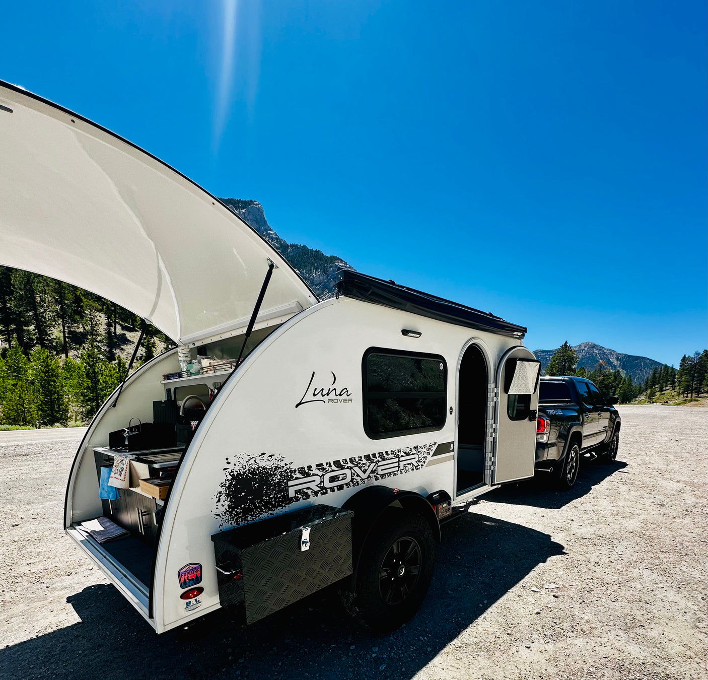 White Luna Rover teardrop RV rental with open galley, hitched to a truck, in a mountain landscape under a clear blue sky.