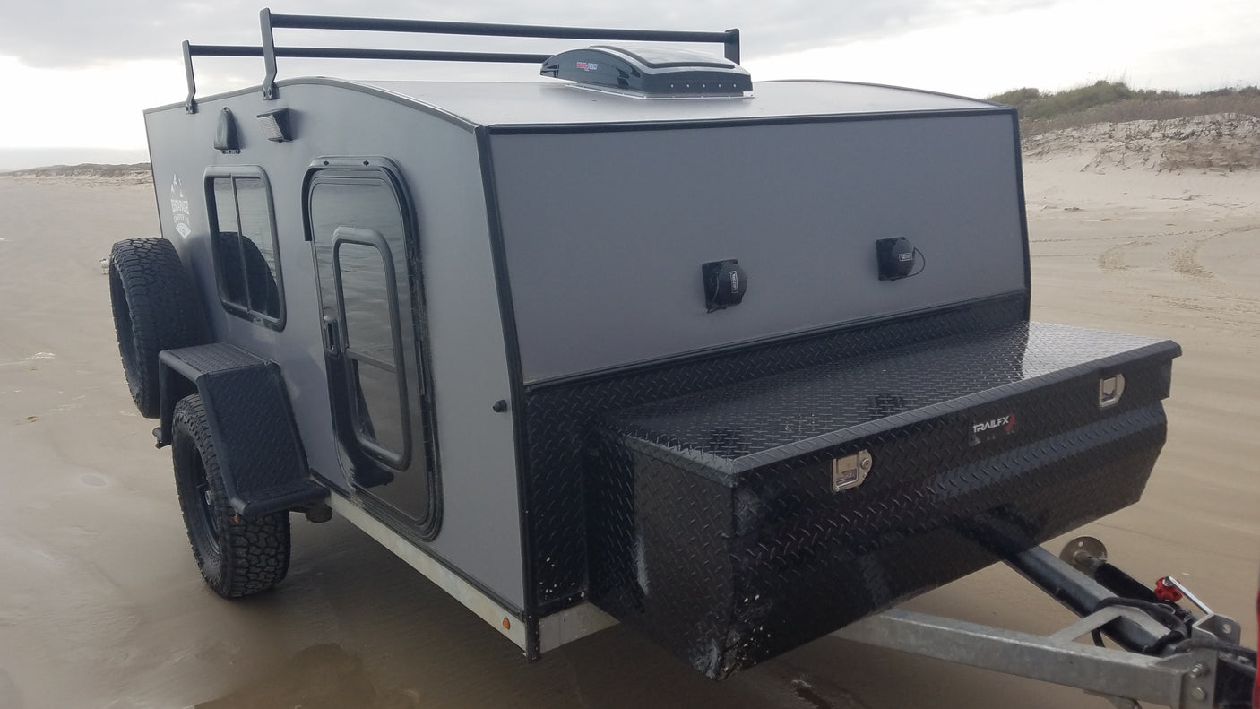 A sleek gray teardrop RV rental on a sandy beach. This compact off-road camper features a roof rack and front storage.