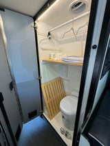 Step inside this compact RV rental wet bath, featuring a toilet, shower mat, storage shelf, and hanging rod.