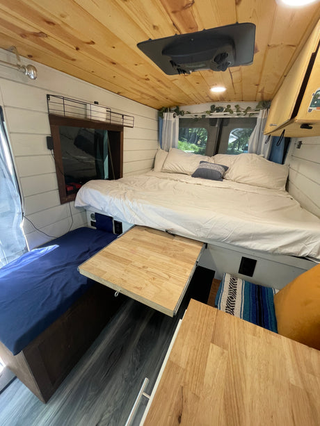 Cozy RV rental interior featuring a large bed, wooden ceiling, white walls, and a pull-out table. Ideal for adventure travel.