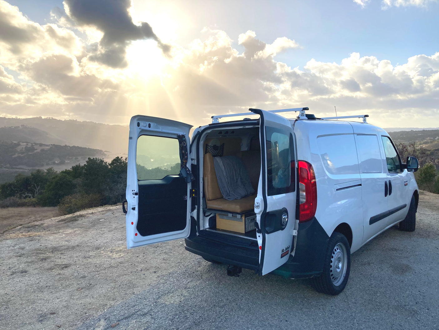 An RV rental camper van with open rear doors revealing its interior, parked on a scenic hill as sunbeams pierce through clouds.