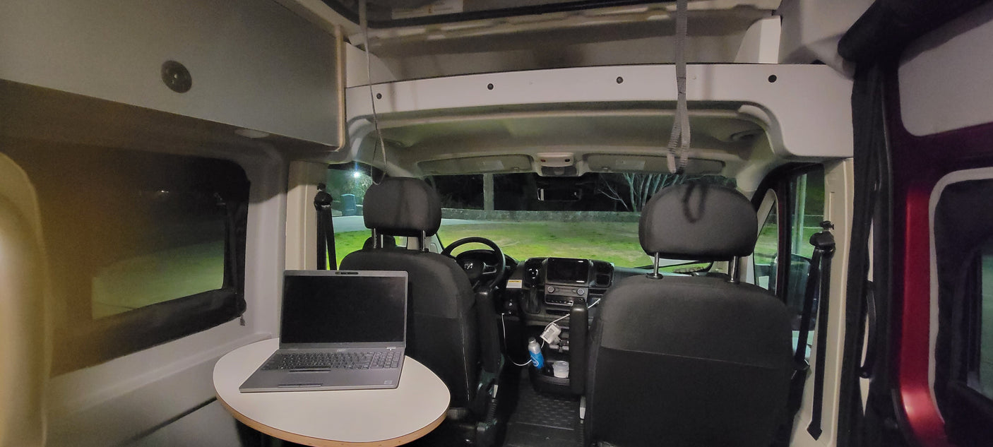 Inside an RV rental at night: a well-lit mobile office space with a laptop on a table, facing the driver's cabin.