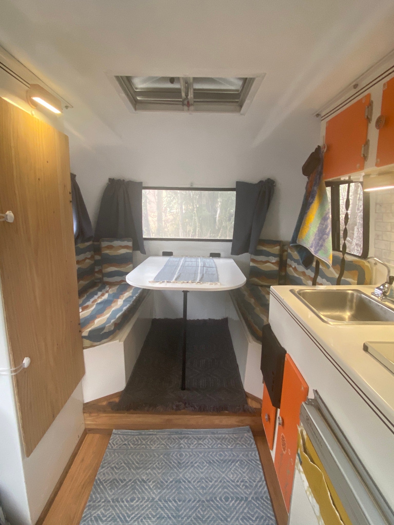 This cozy RV rental interior features a dinette with patterned cushions, a compact kitchen, and a window revealing trees.