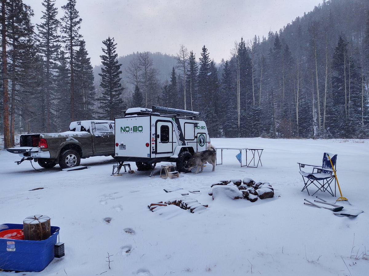 Winter RV rental adventure: A truck and NOBO camper trailer parked in a snowy forest, with a dog, fire pit, and gear. — photo 12