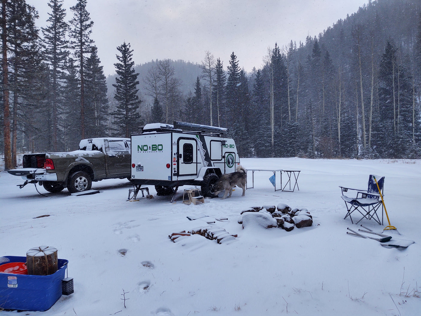 Winter RV rental adventure: A truck and NOBO camper trailer parked in a snowy forest, with a dog, fire pit, and gear.
