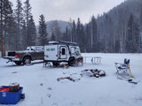Winter RV rental adventure: A truck and NOBO camper trailer parked in a snowy forest, with a dog, fire pit, and gear.