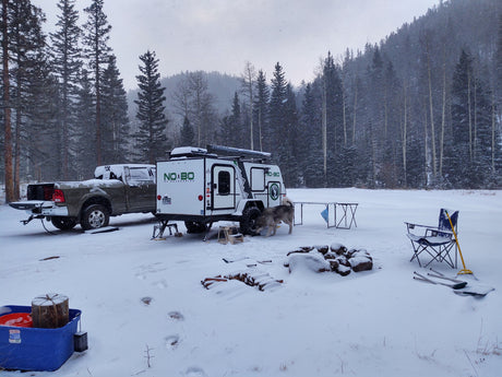 Winter RV rental adventure: A truck and NOBO camper trailer parked in a snowy forest, with a dog, fire pit, and gear.