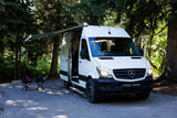 Enjoy nature with this Mercedes Sprinter rv rental camper van. Awning extended, chairs out, nestled among green trees.