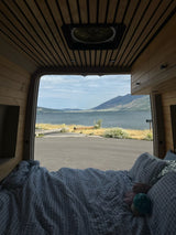 Enjoy the serene view from an RV rental's rear bed, featuring a cozy wood-paneled interior, a lake, mountains, & asphalt road.