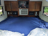 Inside an RV rental: a bed with blue blanket, wooden cabinets, TV, AC, and charging station for comfortable travel.
