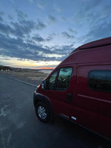 Red RV rental parked roadside at a stunning sunset. Camper van overlooks water with distant hills & lights.