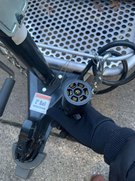 Gloved hand holding a 7-pin trailer connector, preparing an RV rental hitch. Note: 2" ball required for safe towing.