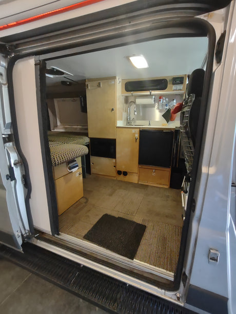 Interior view of an RV rental camper van, showing a compact kitchen with sink and storage, plus a living/sleeping area.