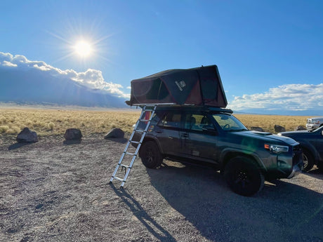 An RV rental SUV with a rooftop tent & ladder set up for camping, with sunny mountains & golden fields in the background.