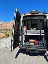 RV rental camper van with open rear, revealing a bed & storage. Parked in a sunny desert landscape with mountains.