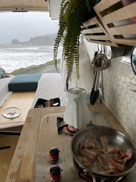 Inside an **rv rental** van, breakfast bacon cooks on the stove beside a boiling kettle, offering a cozy view of the stormy ocean.