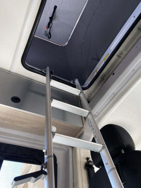 Interior of an RV rental: A silver ladder provides access to an upper sleeping bunk, with a roof hatch and storage units.