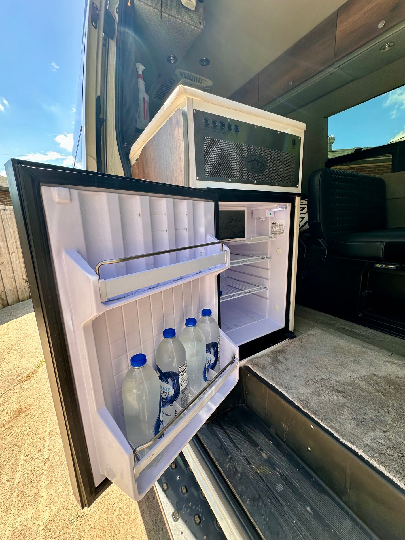 RV rental interior: open fridge with water bottles, a control panel, and seating in a well-equipped camper van.