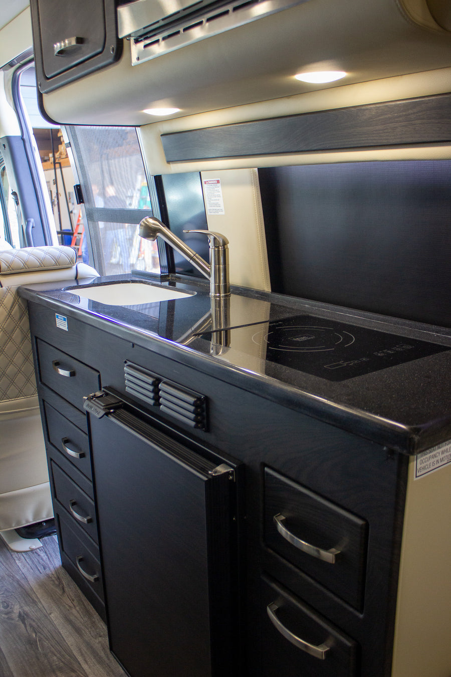 Compact RV rental kitchen featuring a sink, induction stove, fridge, and dark cabinetry.