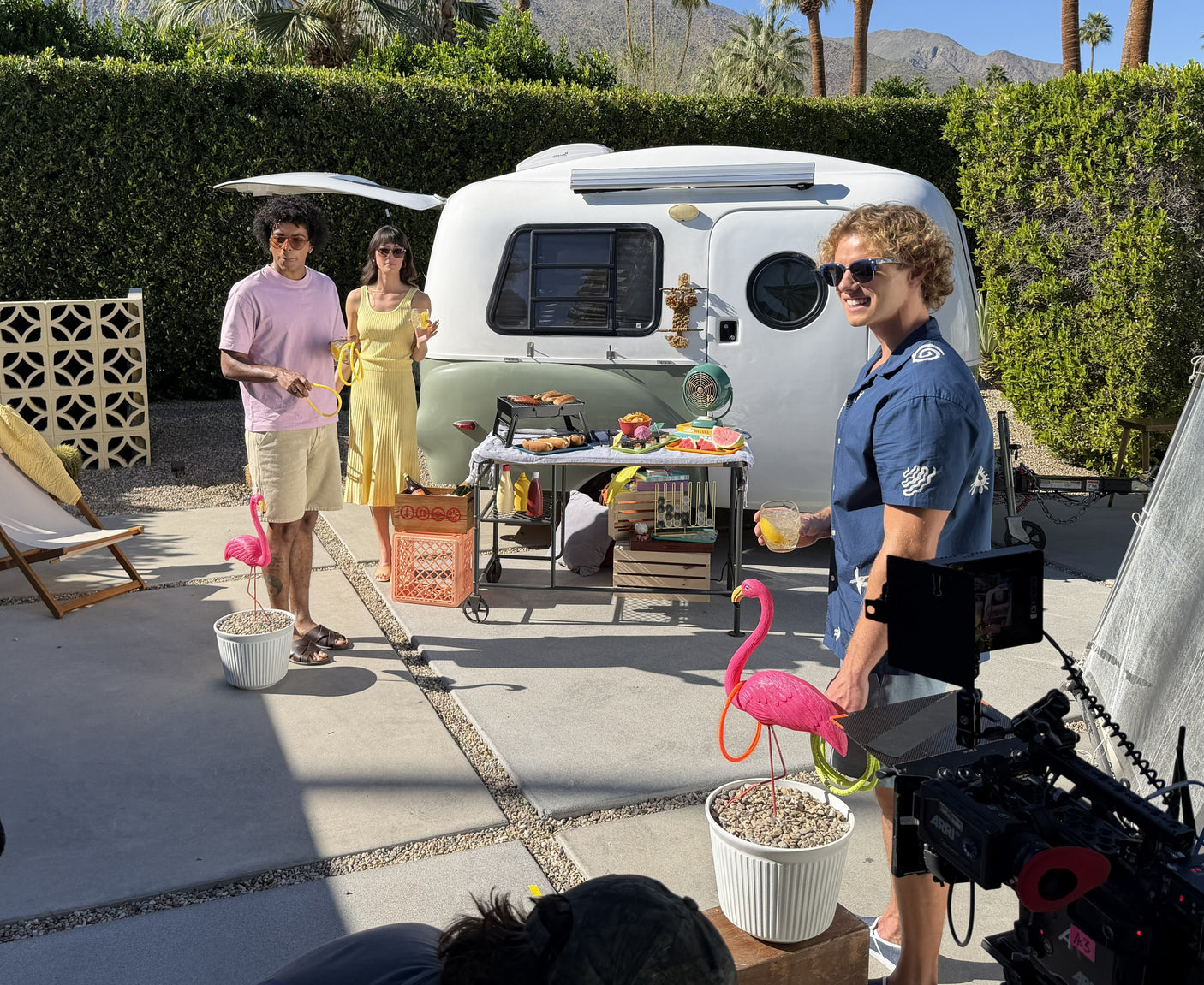 RV rental photoshoot: smiling cast, BBQ, pink flamingos, and a vintage travel trailer in a sunny setting with mountains.