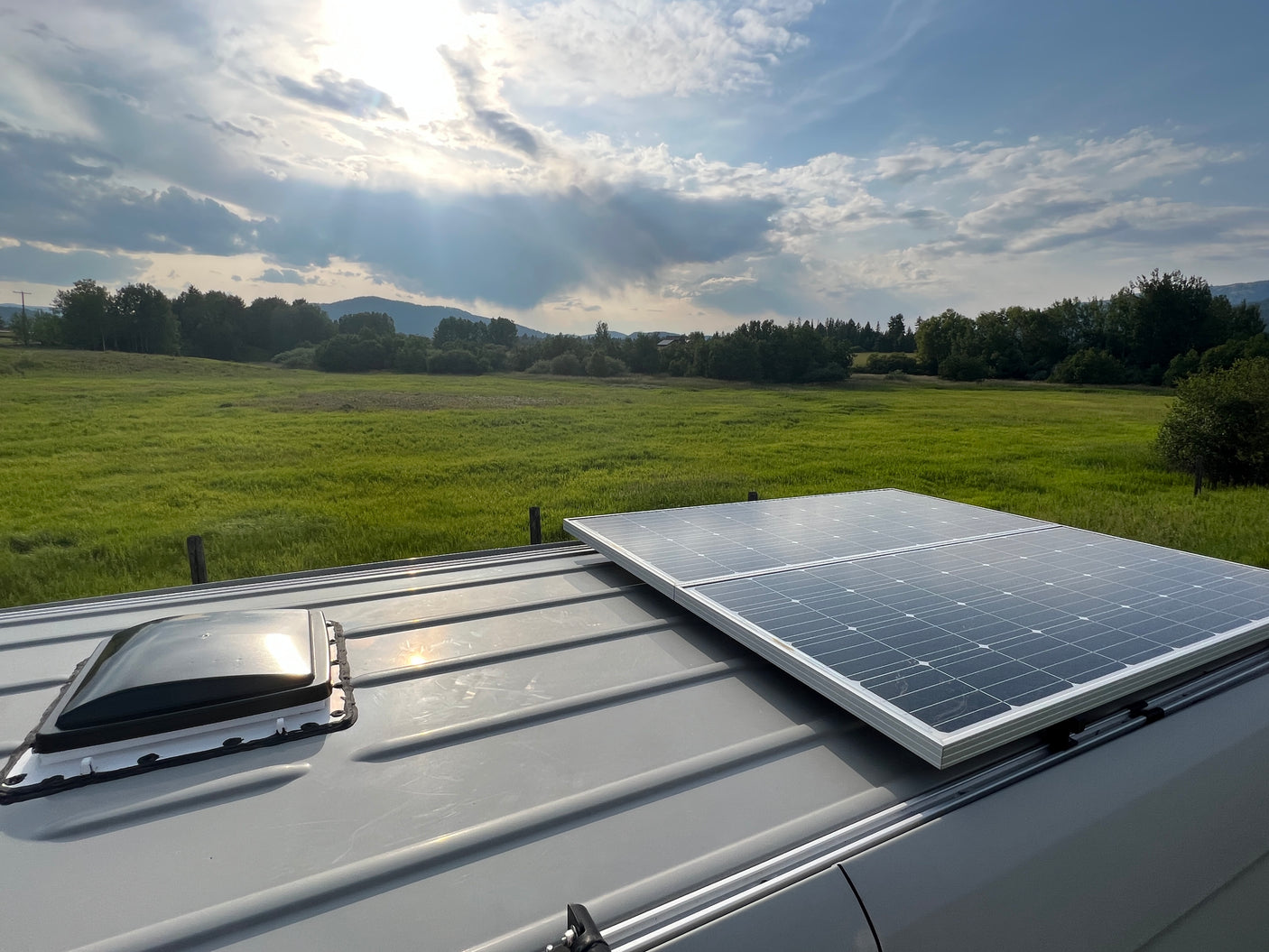 View from an RV rental roof: solar panels, vent, green field, trees, and mountains under a cloudy sky. Ideal for outdoor adventures.