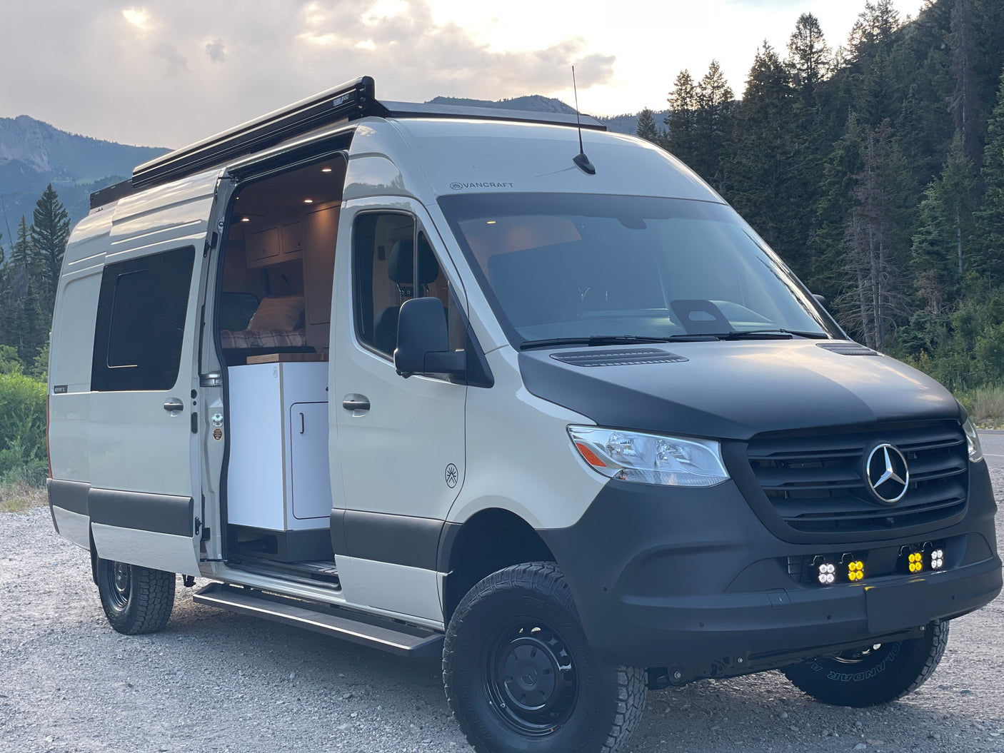 A custom white & black Mercedes-Benz Sprinter camper van rv rental, parked on gravel in a scenic mountain setting.