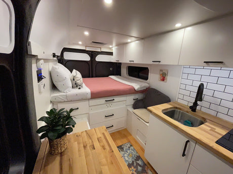 Explore this compact RV rental interior with a cozy bed, sleek kitchen, white cabinets, and clever storage solutions.