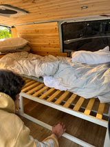 Cozy RV rental bed with wooden slatted frame and a person accessing under-bed storage in a camper van.