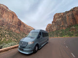 An RV rental van is parked on a scenic road, flanked by towering red rock cliffs under a cloudy sky.