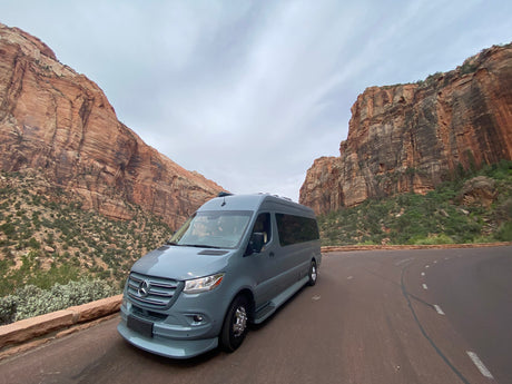 An RV rental van is parked on a scenic road, flanked by towering red rock cliffs under a cloudy sky.