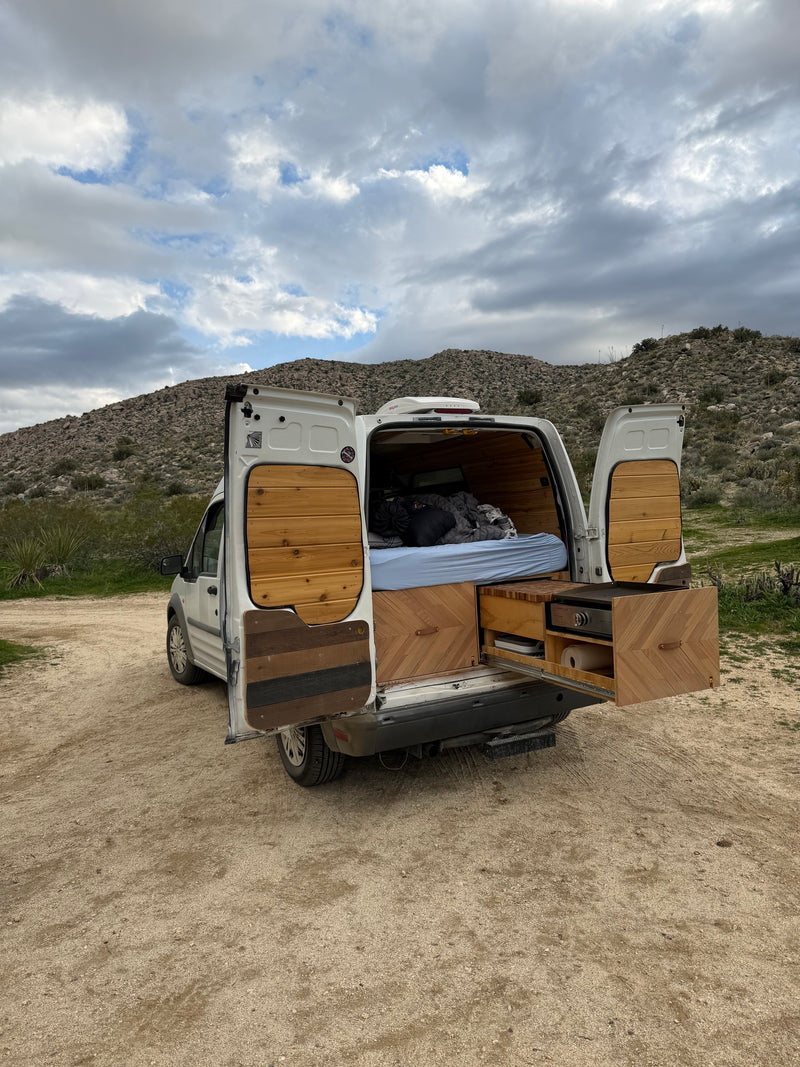 This RV rental shows a custom camper van with open back, revealing a comfy bed & pull-out kitchen in a desert setting. — photo 7