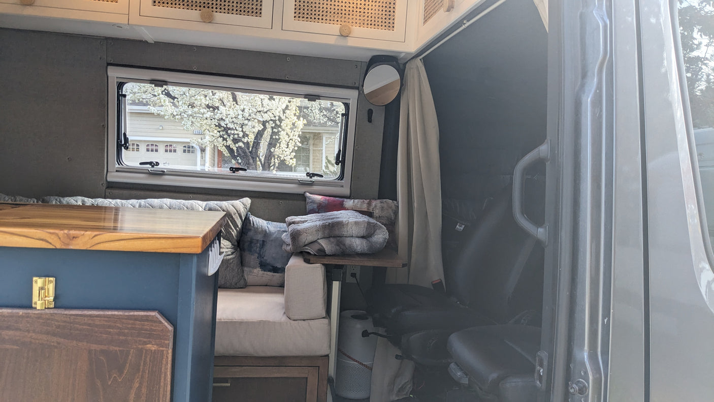 Cozy RV rental interior with a window view of a blossoming tree, comfy seating, wooden finishes, and a small mirror.