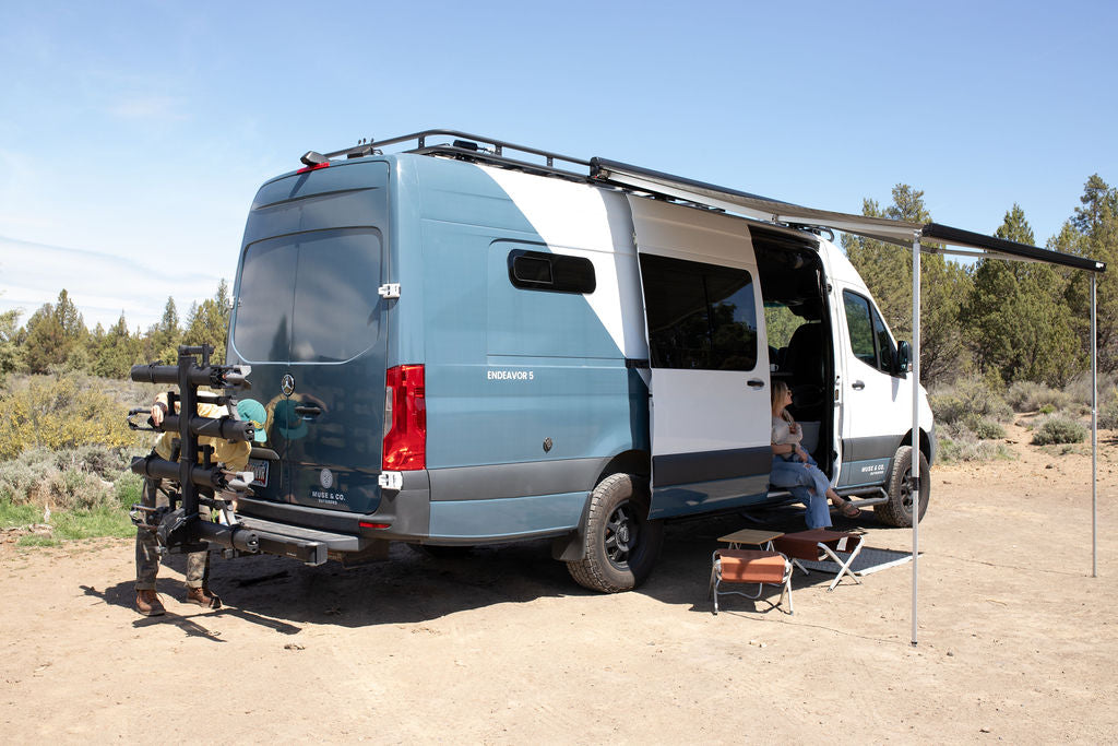 An RV rental camper van parked in nature. A person relaxes under the awning, another handles the bike rack at the back.